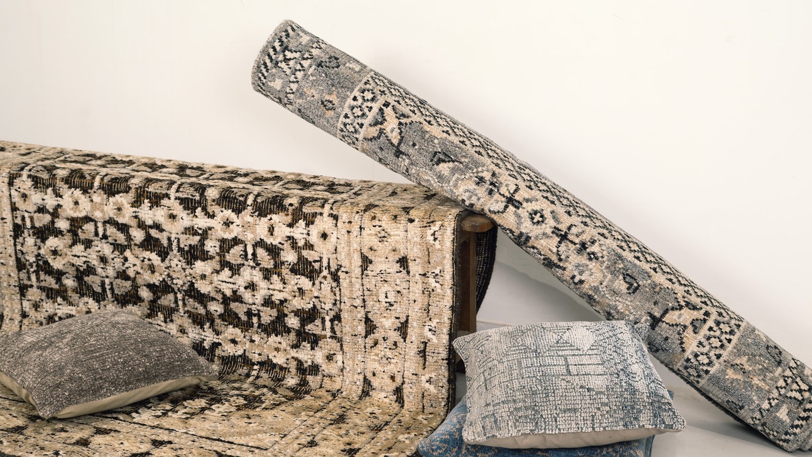 A rolled patterned rug and a folded rug beside a cushion against a plain background