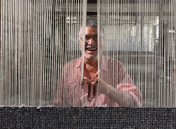 A weaver seated behind the warp threads of a loom, hands raised at the work