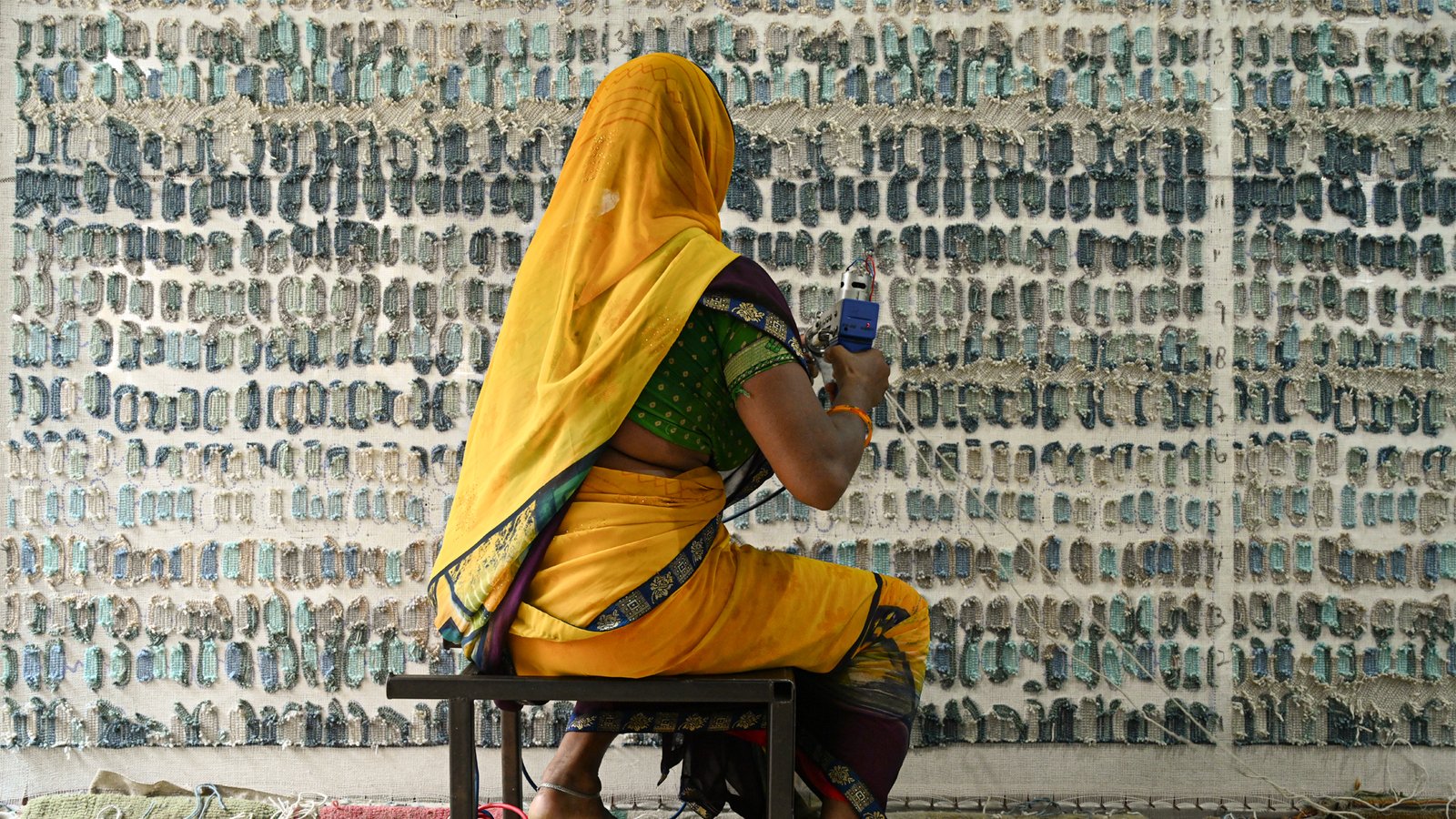A woman in a yellow sari seated at a loom, working on a large carpet in progress