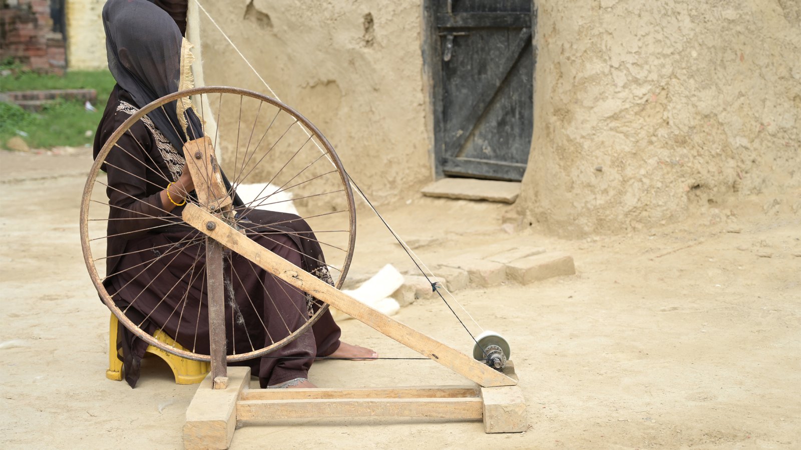 A woman spinning yarn at a traditional charkha outside a mud-walled house